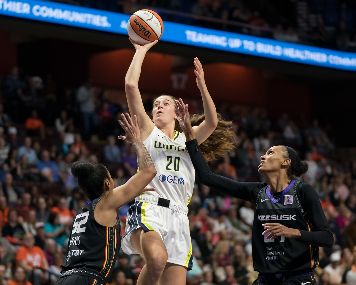 Dallas Wings combo forward Maddy Siegrist rises midair with the ball in her hand to shoot over Connecticut guard Tyasha Harris while Sun forward DeWanna Bonner stands next to them with a raised arm trying to help contest the shot. Behind the three of them, a packed crowd in the stands for this WNBA game.