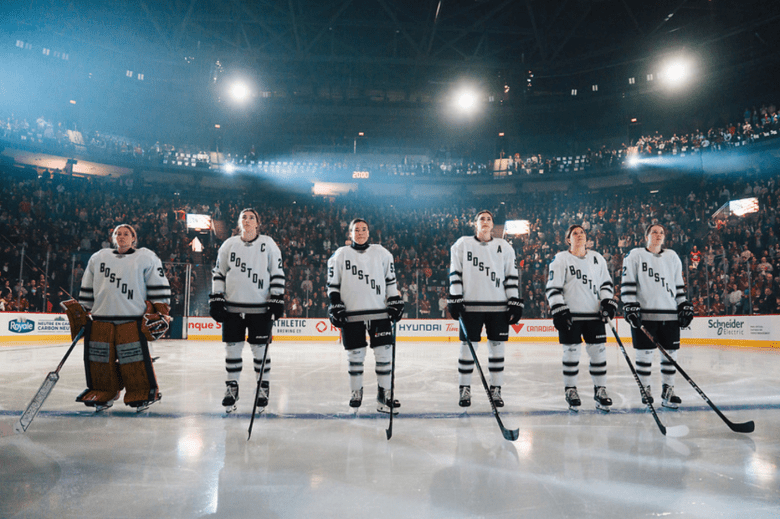 Five Boston players stand on the blue line with their helmets off. They're wearing white away uniforms.