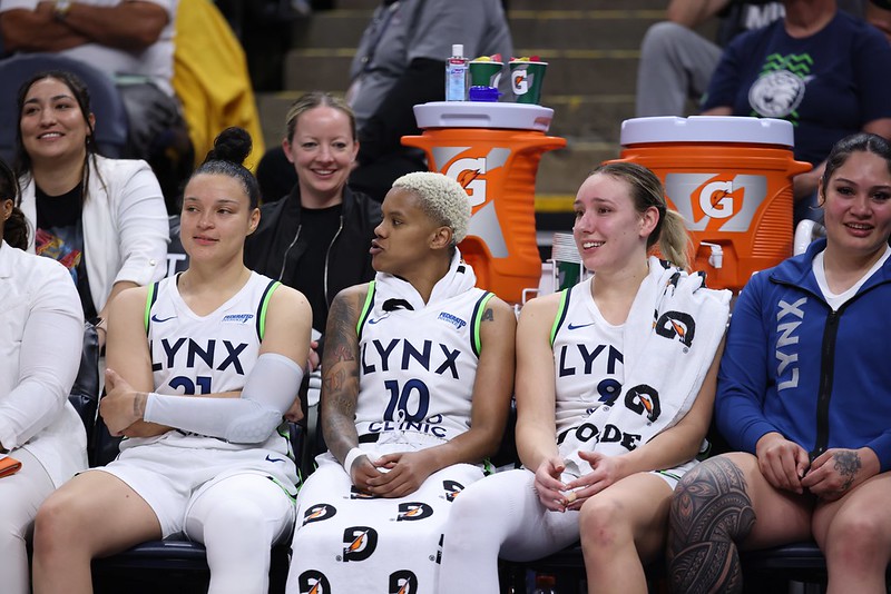 Minnesota Lynx players sit on the bench wearing white uniforms. Assistant athletic trainer Brandi BlueArm and sports performance coach Andrea Hayden sit behind them next to large orange Gatorade containers.