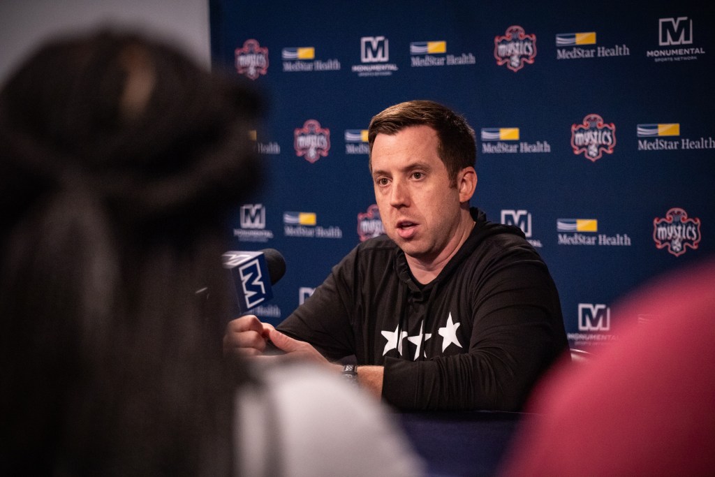 Washington Mystics head coach Eric Thibault sits at the podium to speak with reporters before a game. Behind him is a blue backdrop with Mystics, Monumental Sports Network and MedStar Health logos.