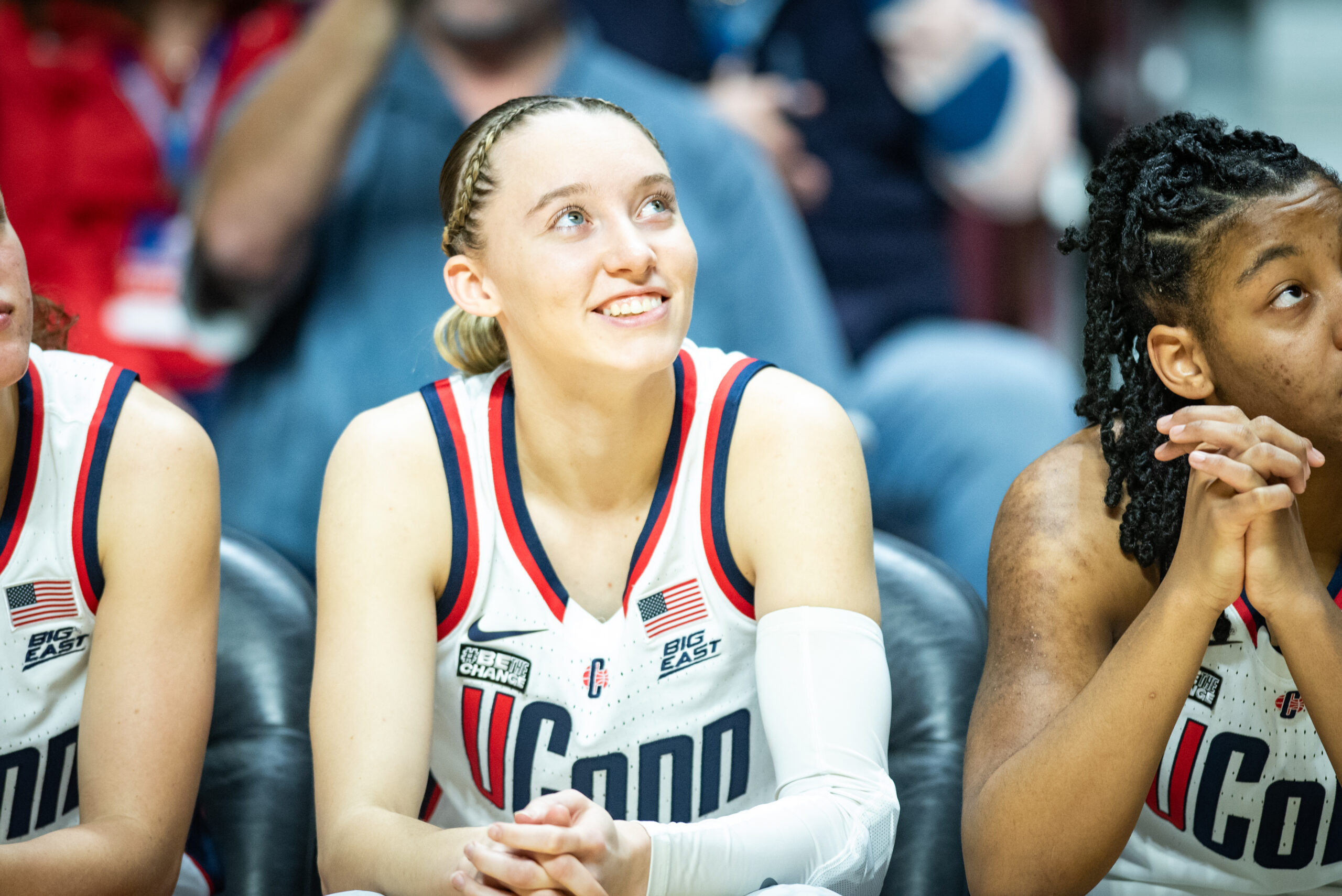 Paige Bueckers is seated on the bench wearing a UConn home white jersey, looking up and smiling
