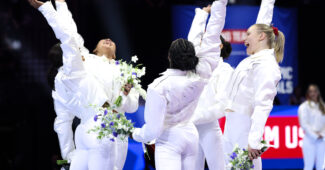 Five gymnasts in white warmup suits celebrate with their hands raised.