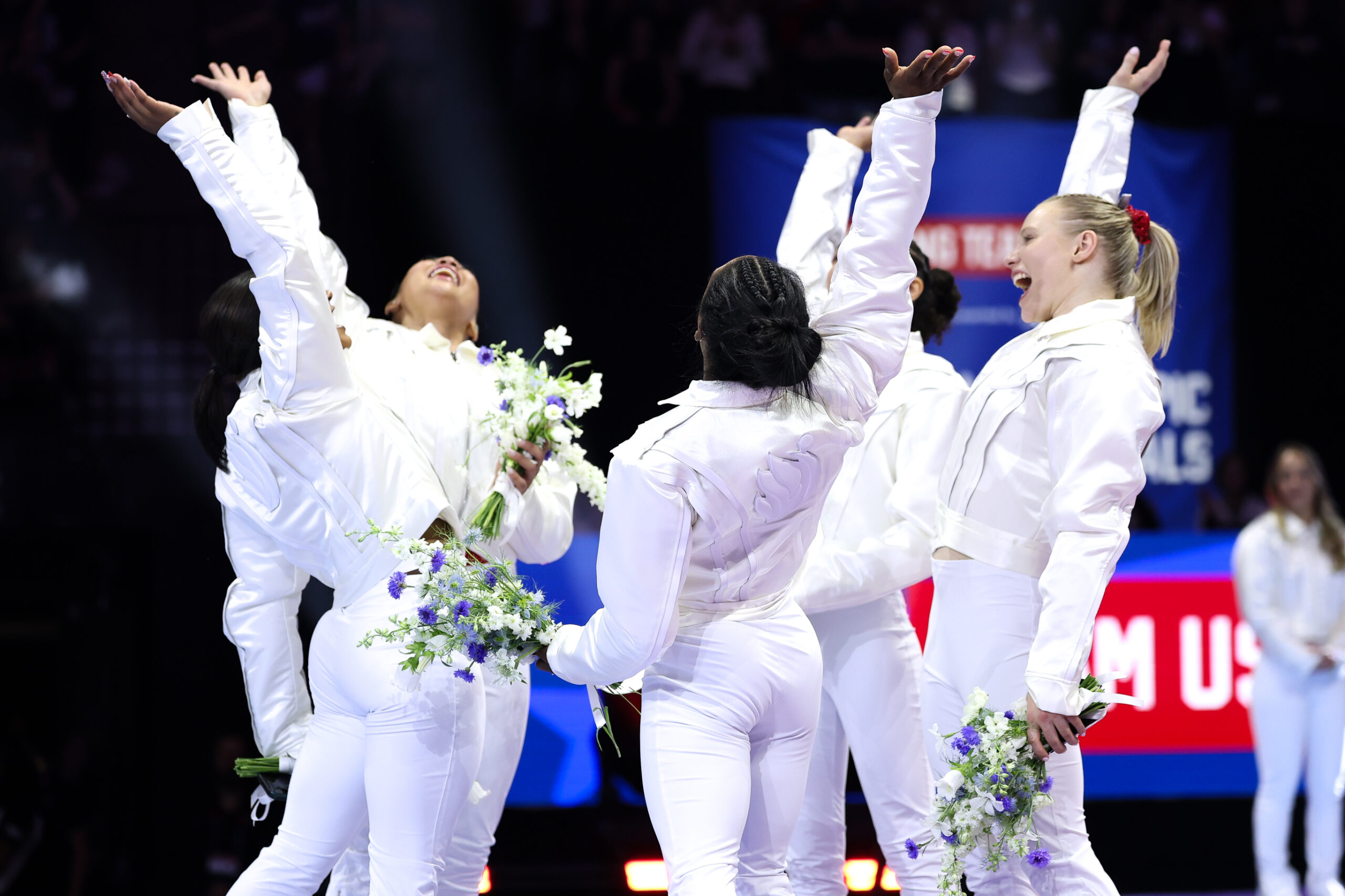 Five gymnasts in white warmup suits celebrate with their hands raised.