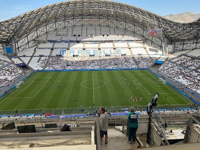 Inside of Marseille Olympic soccer field is pictured with a small crowd of fans in the stands. The work "Marseille" can be seen in the stands in blue text on white seats.
