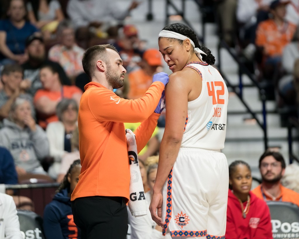 Connecticut Sun head athletic trainer Ryan Newton wears an orange jacket, black pants and blue rubber gloves. He holds a white towel with Gatorade logos on it and examines Sun forward Brionna Jones' left shoulder on the sideline.