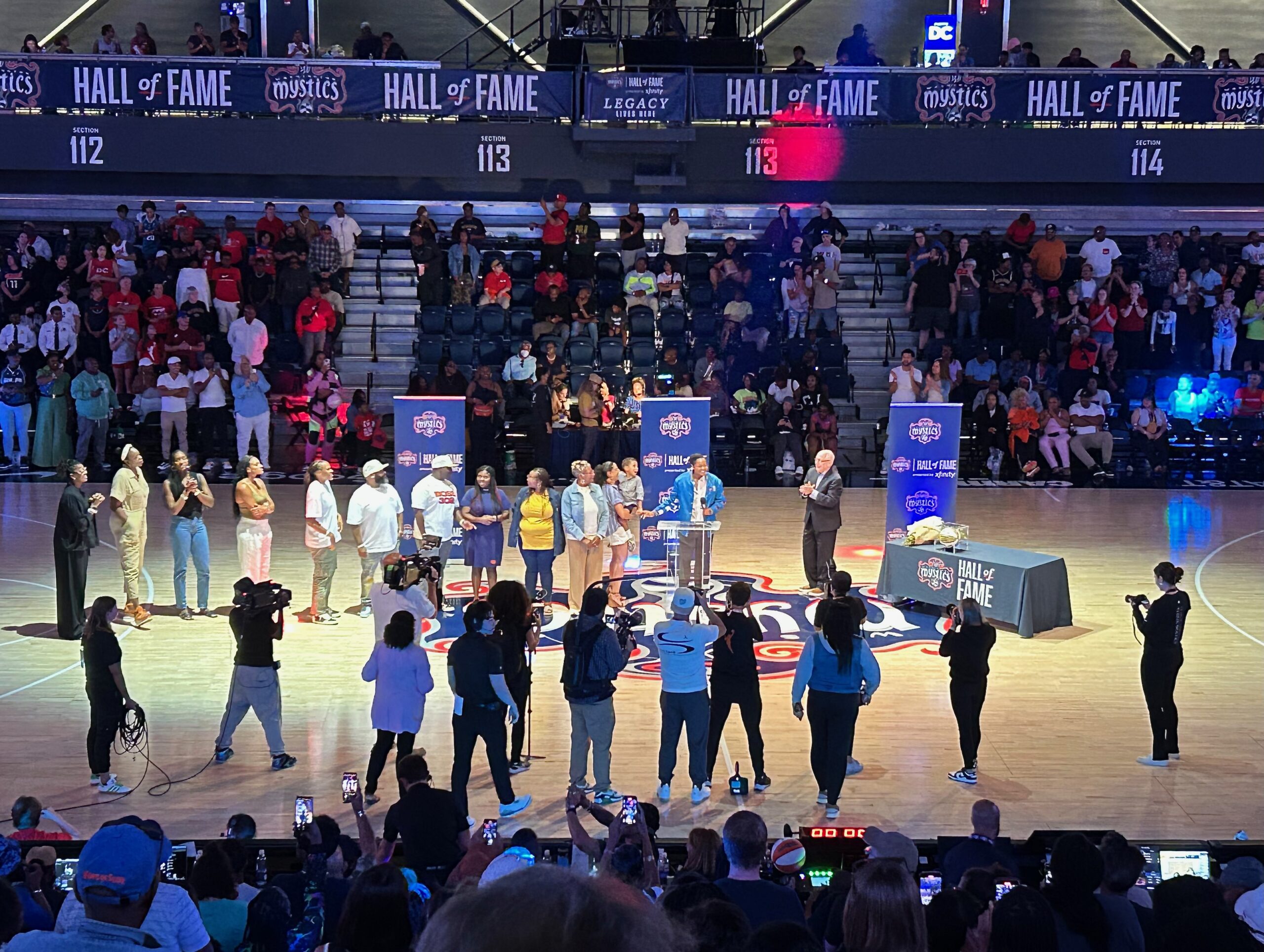 Former Washington Mystics guard/forward Alana Beard stands at a glass podium at center court and addresses the crowd during her Hall of Fame induction. Mystics general manager Mike Thibault and Beard's family and friends stand on either side of her.