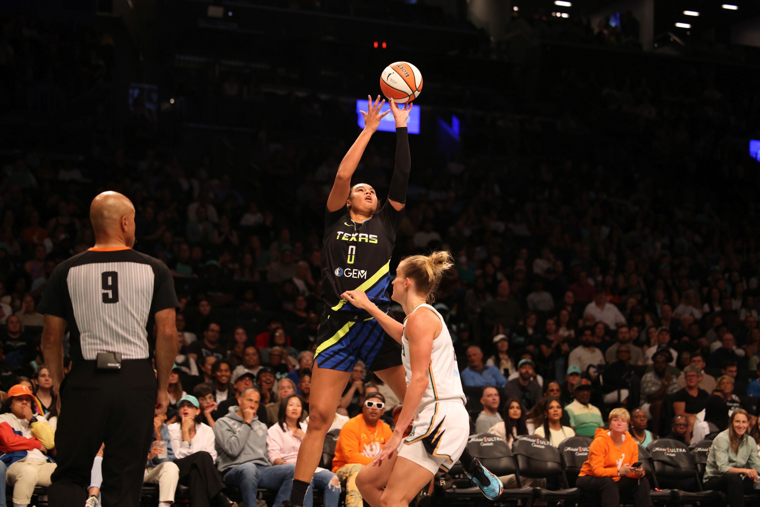 Dallas Wings forward Satou Sabally releases a jump shot off her fingertips in midair as New York Liberty off-ball guard Leonie Fiebich stands in front of her on defense, while a packed WNBA crowd watches from behind