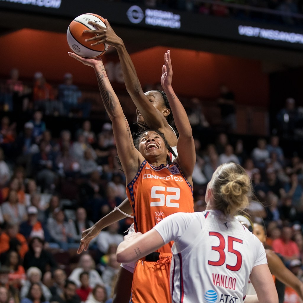 Positioned behind Connecticut Sun guard Tyasha Harris, Washington Mystics guard Shatori Walker-Kimbrough jumps and reaches with her left hand to block Harris' shot. Walker-Kimbrough has her whole hand on the side of the ball, which is just leaving Harris' right palm.