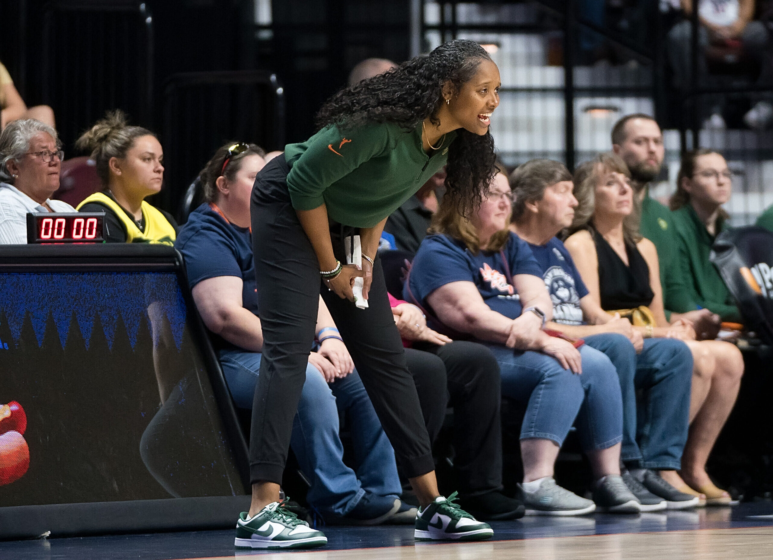 Seattle Storm head coach Noelle Quinn bends over in a wide stance, stat sheet in hand, and relays information to her players from the sideline.