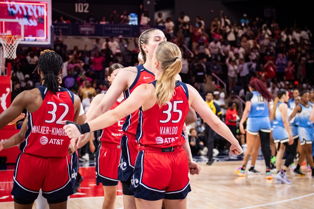 Washington Mystics guard Julie Vanloo and forward Emily Engstler chest-bump to celebrate a win. Other Mystics players gather nearby, and several members of the losing Chicago Sky team gather far in the background.