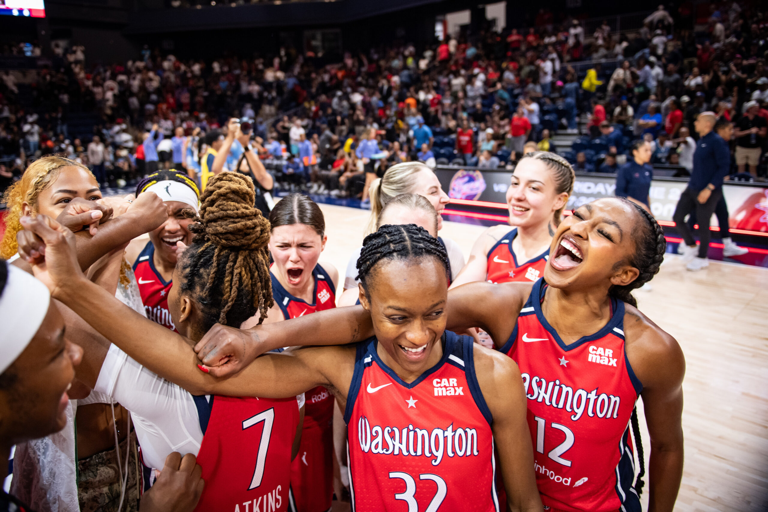 Washington Mystics players stand near midcourt after a game and put their fists together in a huddle. The players are each smiling or shouting in celebration.