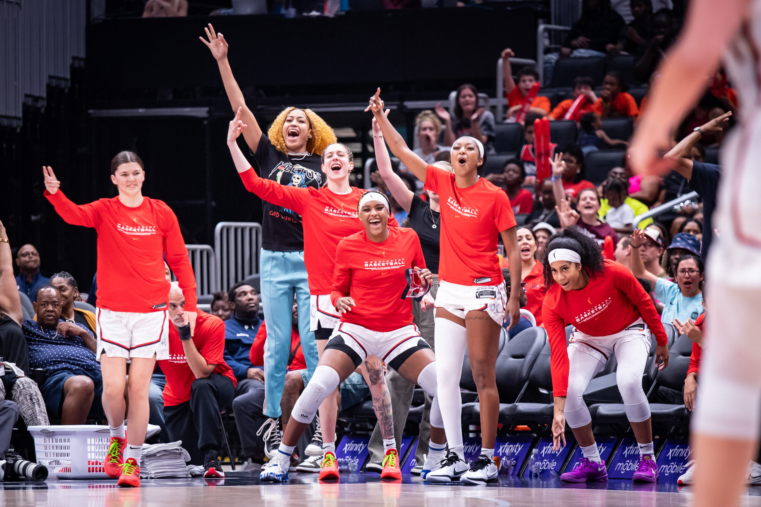 Several Washington Mystics players who are on the bench celebrate a play by shouting, making 3-point signs with their fingers and putting their hands in the air.