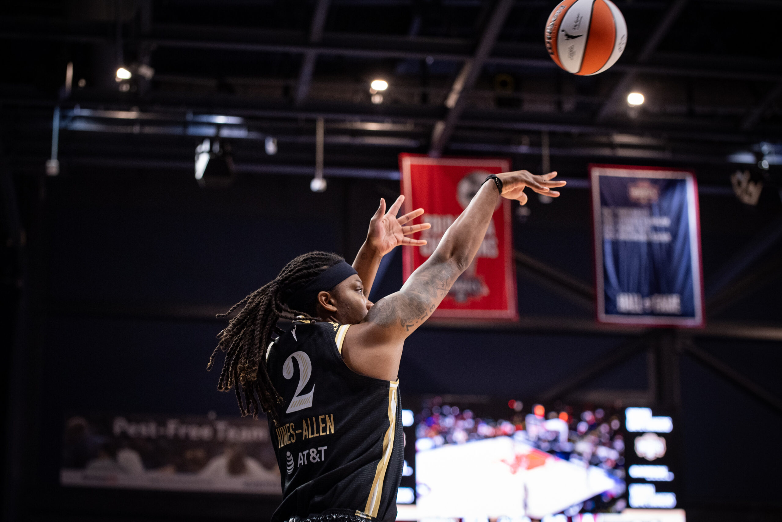 A close-up photo of Washington Mystics forward Myisha Hines-Allen releasing a jump shot. The Mystics' video board and championship banner are visible in the background.