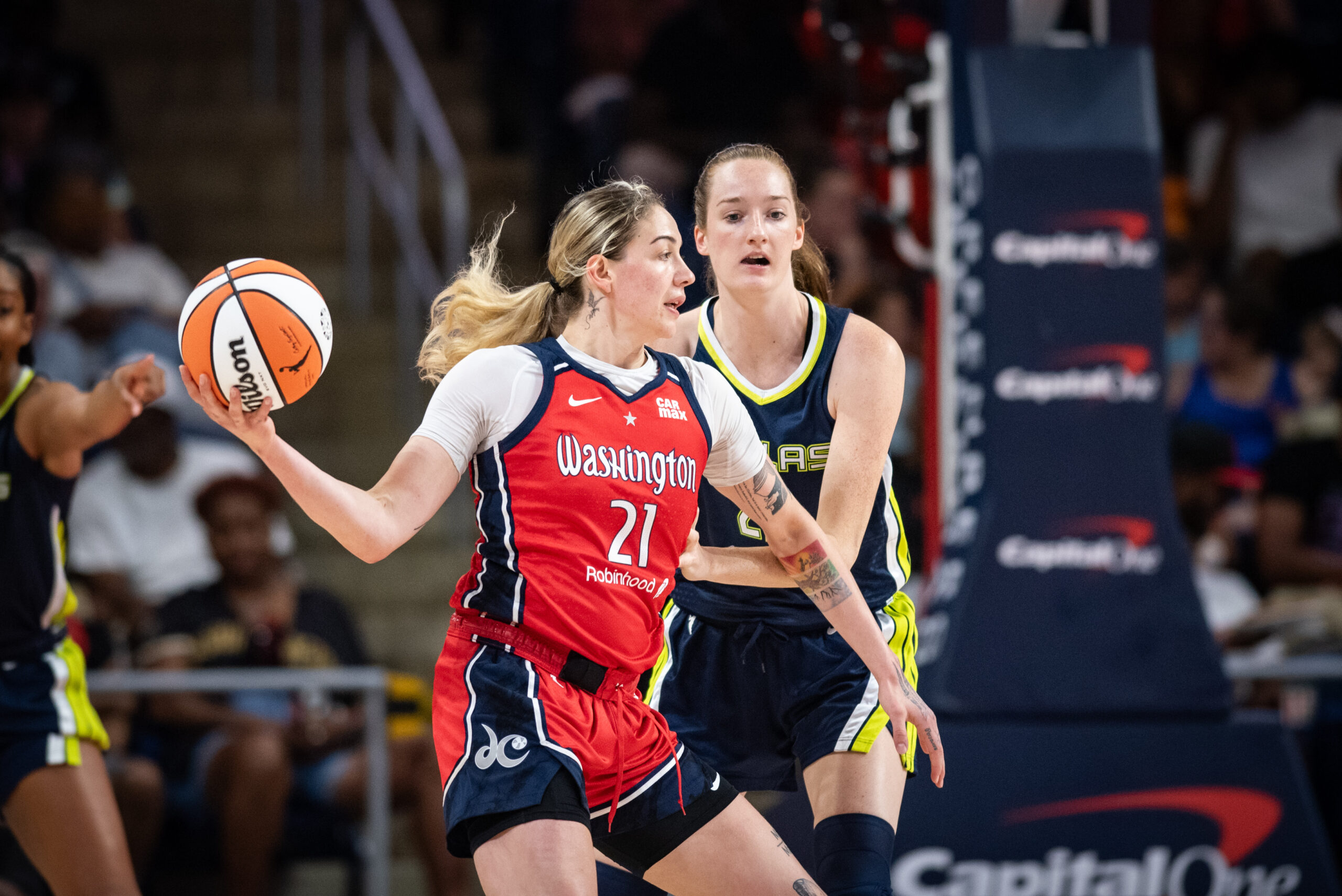 Washington Mystics forward Emily Engstler holds the ball in her hand out to her side as she looks off-camera for an opportunity to pass, as a Dallas Wings defender stands to her other side in a WNBA game with fans out of focus in the background