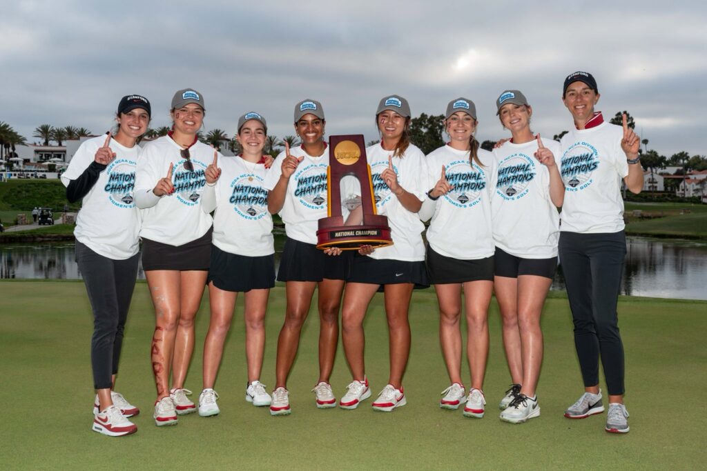 Members and coaches of the Stanford women's golf team pose, each holding up a number one with their fingers, with the NCAA National Championship held in the middle
