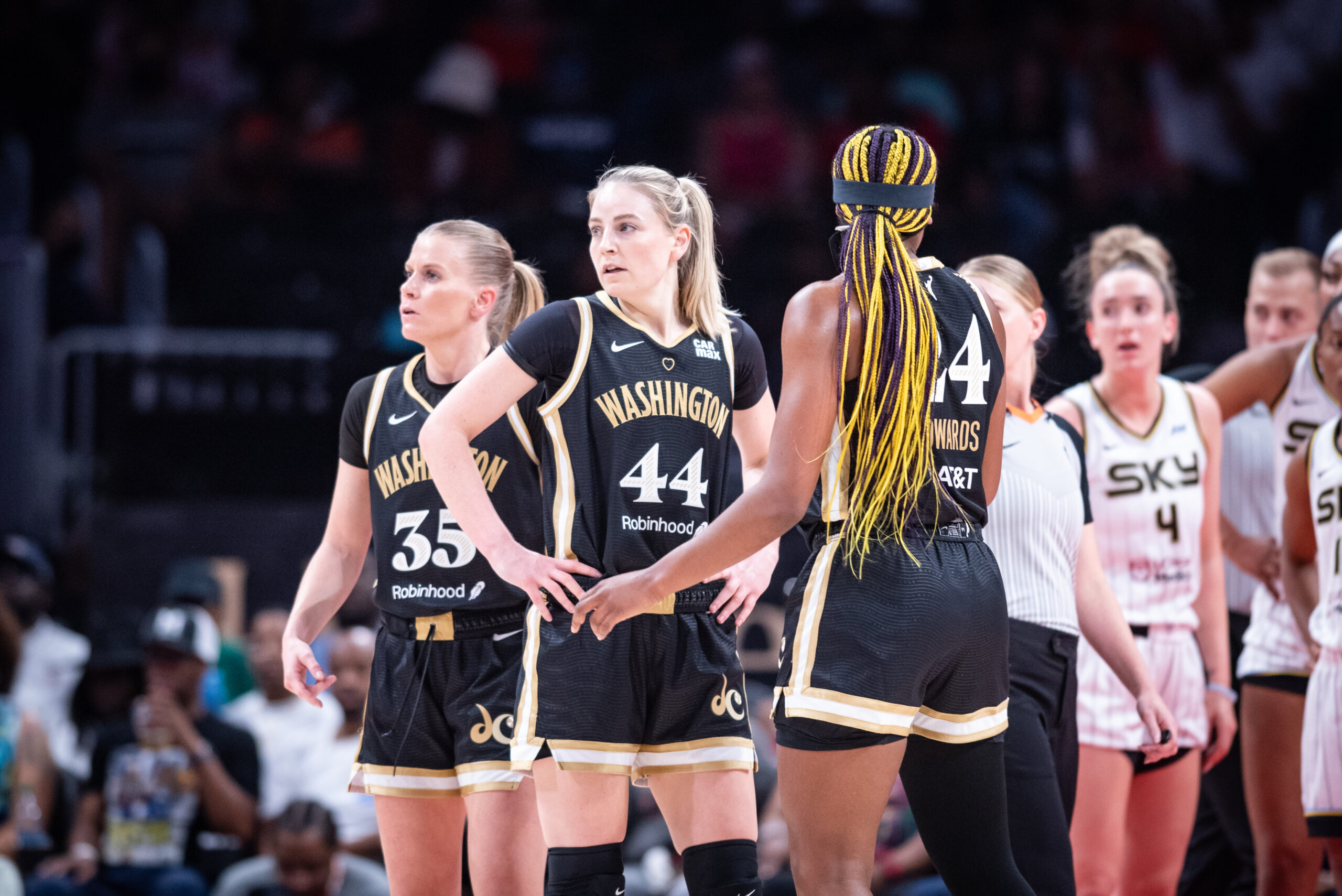 A WNBA game with three players from the Washington Mystics stand together, two looking toward one side of the court and one looking the other way, while a ref and three members of the Chicago Sky stand behind them looking off the court.