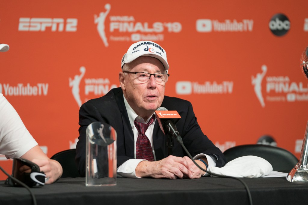 Then-Washington Mystics head coach Mike Thibault sits at the podium wearing a 2019 WNBA champions hat and answering questions from reporters.