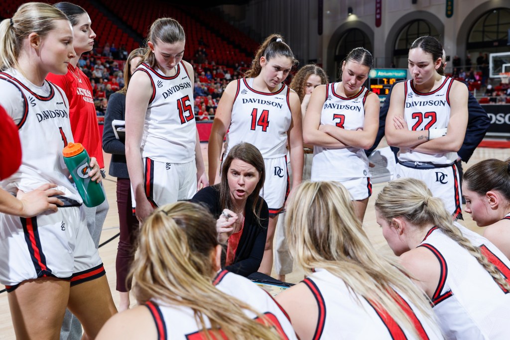 Davidson head coach talks to her team as they listen intently and look at her. The team is gathered in a circle with players standing behind her and players sitting in front of her.