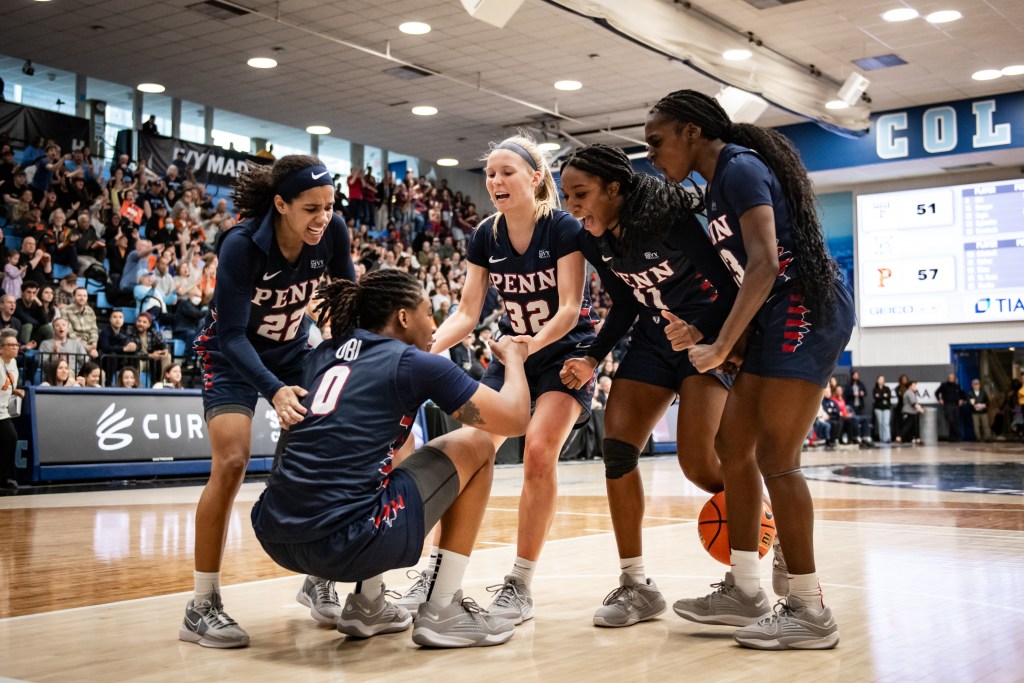 Penn guard Lizzy Groetsch uses both hands to pull forward Jordan Obi to her feet. Guard Mataya Gayle has one hand on Obi's back, and guards Simone Sawyer and Ese Ogbevire are leaning toward Obi and celebrating.