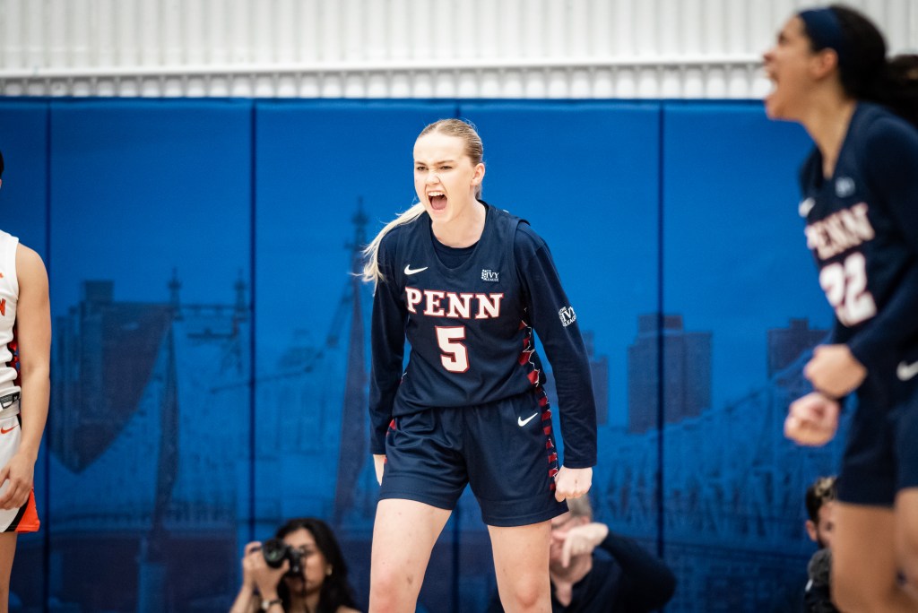 Penn guard Stina Almqvist is shown facing the camera. She screams in celebration, her eyes narrowed. Out of focus in the corner of the frame, her teammate Mataya Gayle is shown in profile, yelling and clenching her fists.
Ivy League