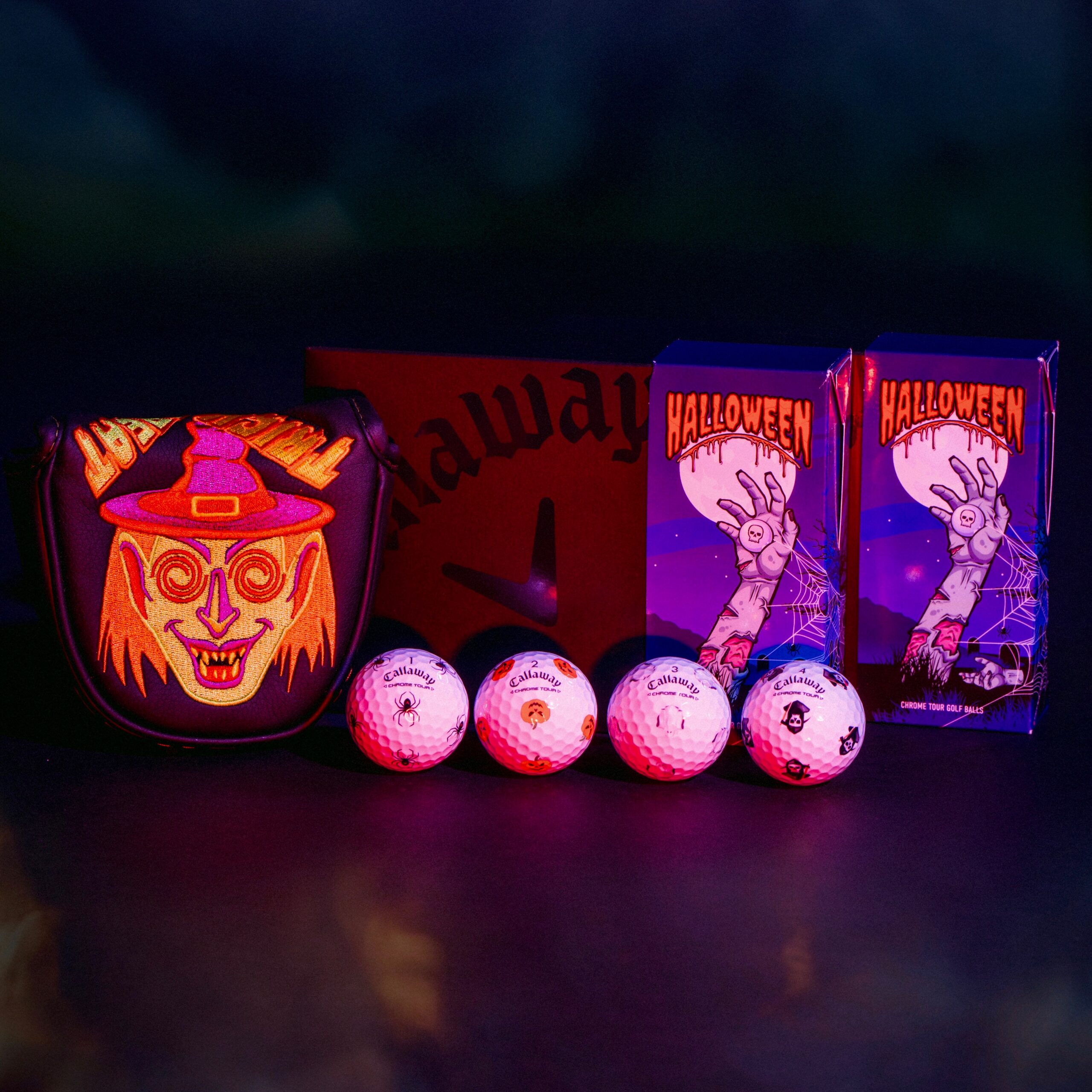 A series of Halloween inspired golf accessories line up across a black background. The items, a putter cover with a witches face and golf balls with spooky icons, seem to glow in the dark.
