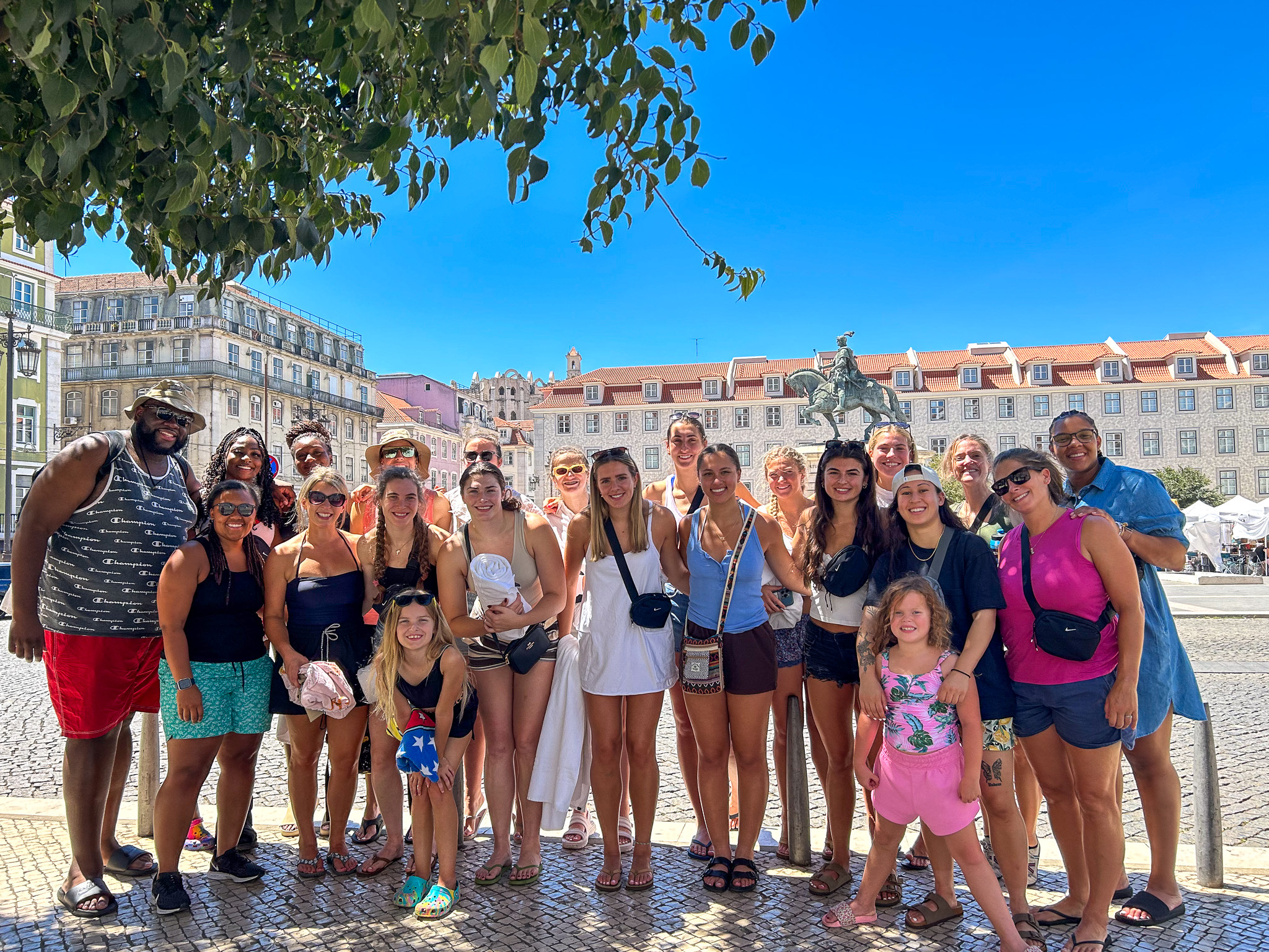 Brown players, coaches, staff and family members smile for a photo on a plaza in Portugal. A statue of someone riding a horse is visible behind them.