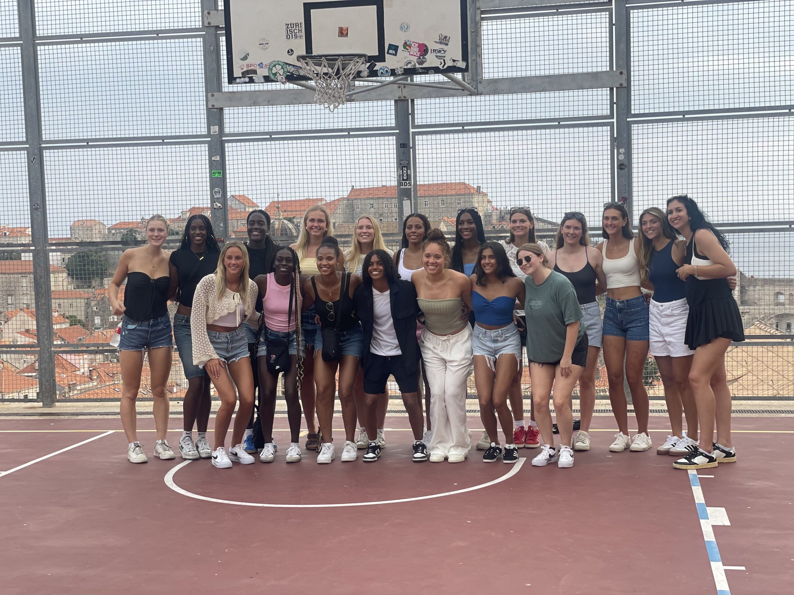 Penn players stand in two rows on an outdoor basketball court, with rooftops visible behind them. They put their arms around each other and smile for a photo.