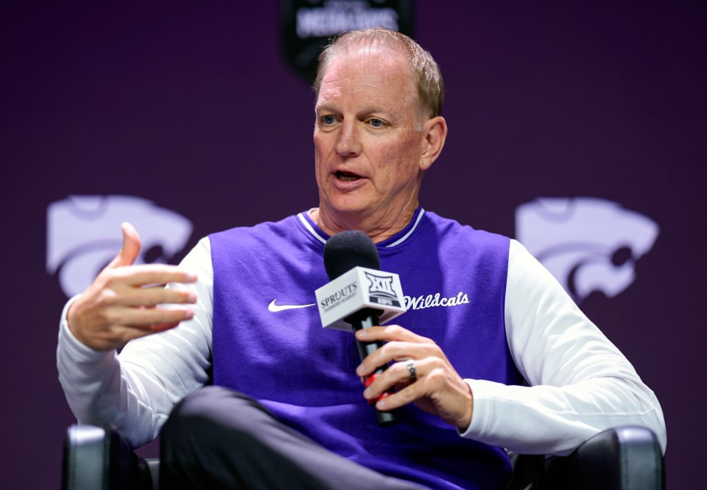 Kansas State head coach Jeff Mittie is seated and speaks into a handheld microphone, gesturing with his free hand.