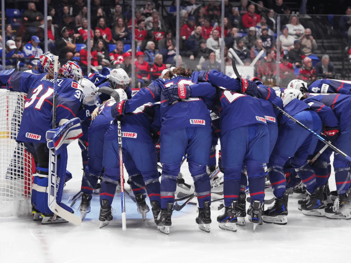 All members of Team USA are arm-in-arm, hunched over in a circle in front of their goal. They are wearing blue uniforms.