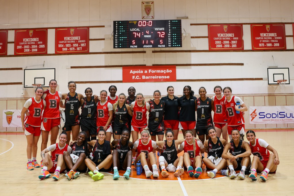 Players from Brown and Lisbon Select pose for a photo with their arms around each other. About half the players are seated on the court, and the rest are standing behind them.