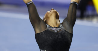 A gymnast in a black leotard caught in a finishing pose on the floor exercise with her arms above her head.