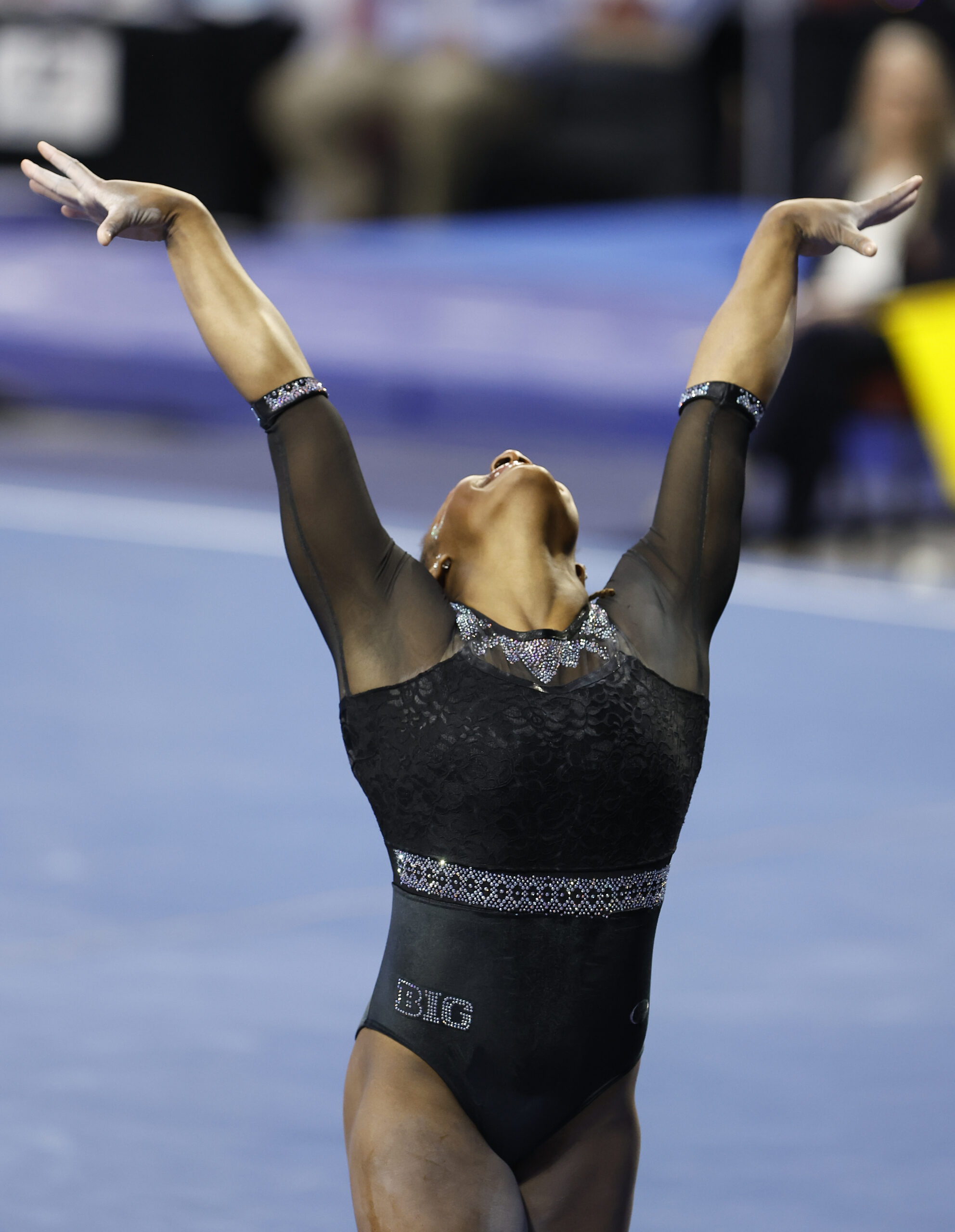 A gymnast in a black leotard caught in a finishing pose on the floor exercise with her arms above her head.