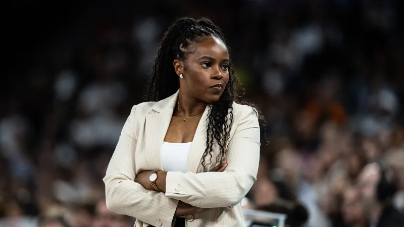 Rena Wakama stands on the sidelines during a game at the 2024 Paris Olympics. She wears a white suit, a crowd watches on from behind her.