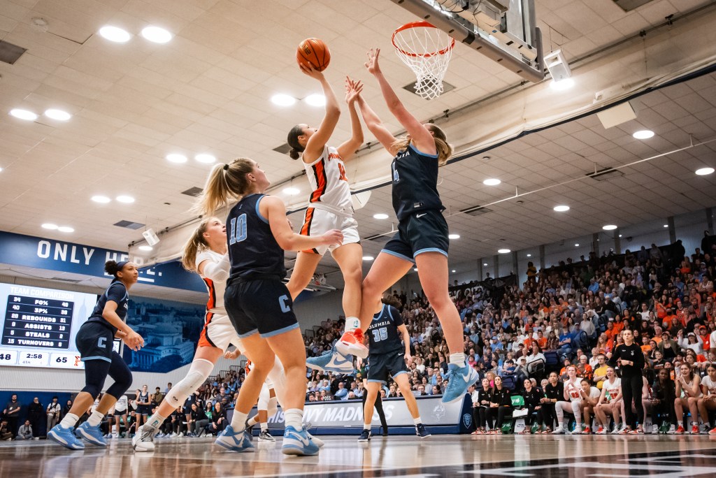 Princeton guard Skye Belker and Columbia guard Fliss Henderson are shown in midair. Belker is trying to shoot a right-handed layup, and Henderson has both hands up to contest it.