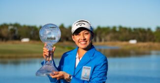 Jeeno Thitikul, the winner of the 2024 CME Group Tour Championship, wears the blue prize jacket and holds up the tournament trophy to her right. She stands in front of the water at Tiburon Golf Club in Naples.