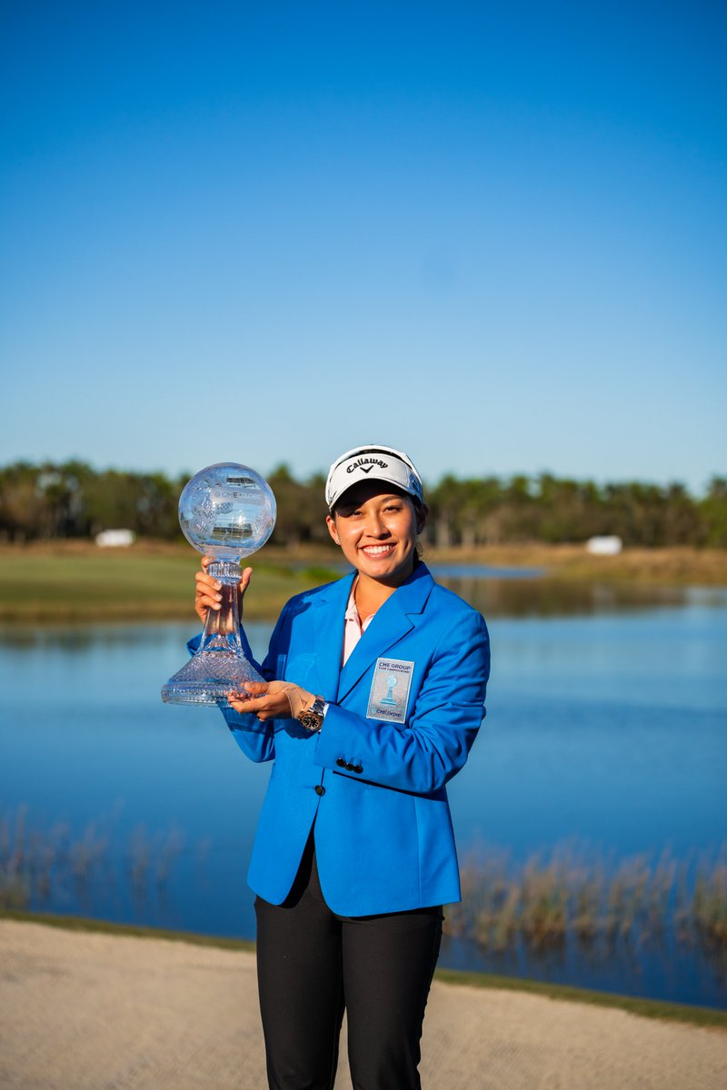 Jeeno Thitikul, the winner of the 2024 CME Group Tour Championship, wears the blue prize jacket and holds up the tournament trophy to her right. She stands in front of the water at Tiburon Golf Club in Naples.