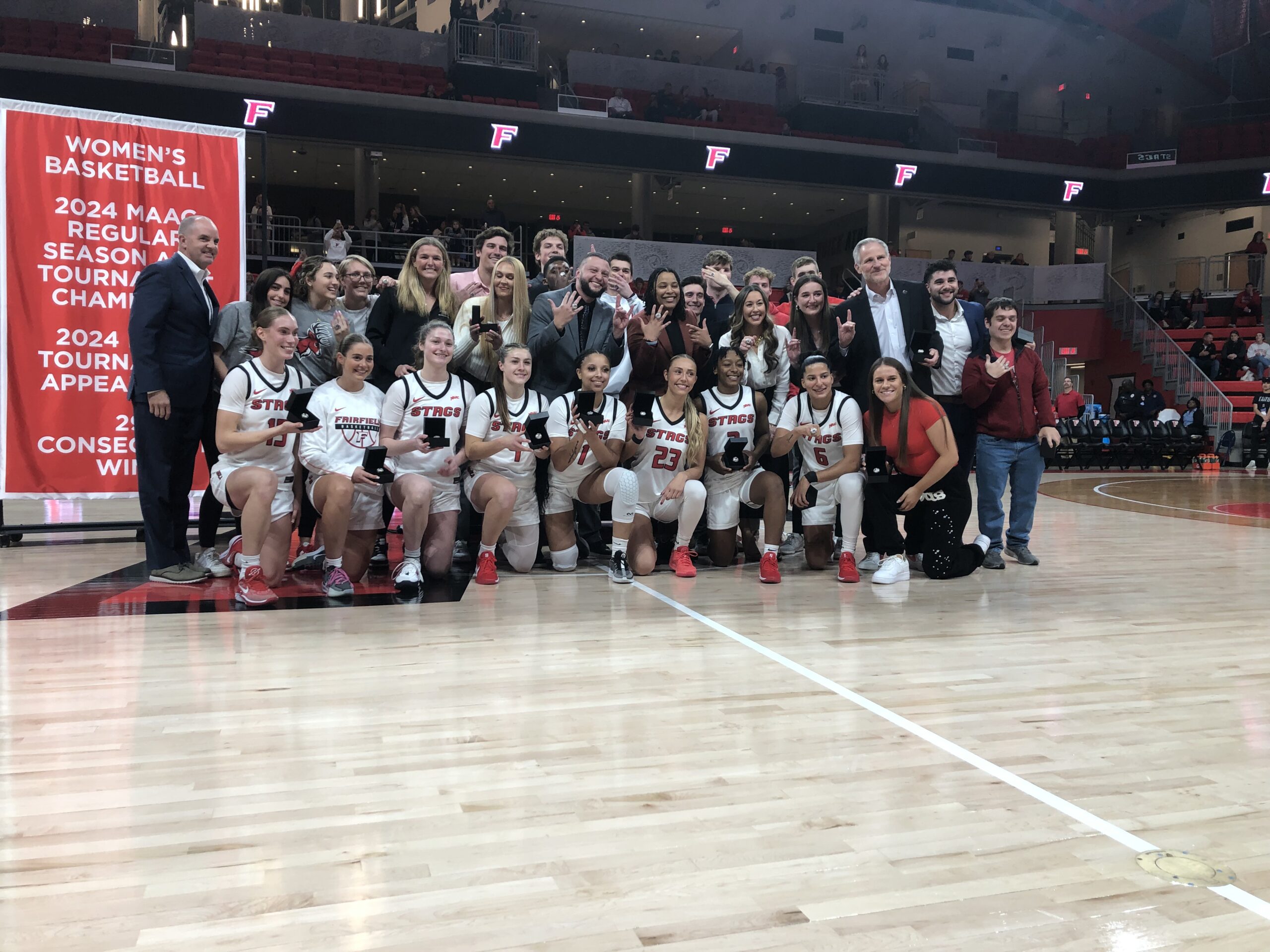 In white and the front row, Fairfield Stags players smile and hold their MAAC championship rings. Above them, coaches, practice players and team staff do the same. (Photo credit: Benjamin Yeargin | The Next)
