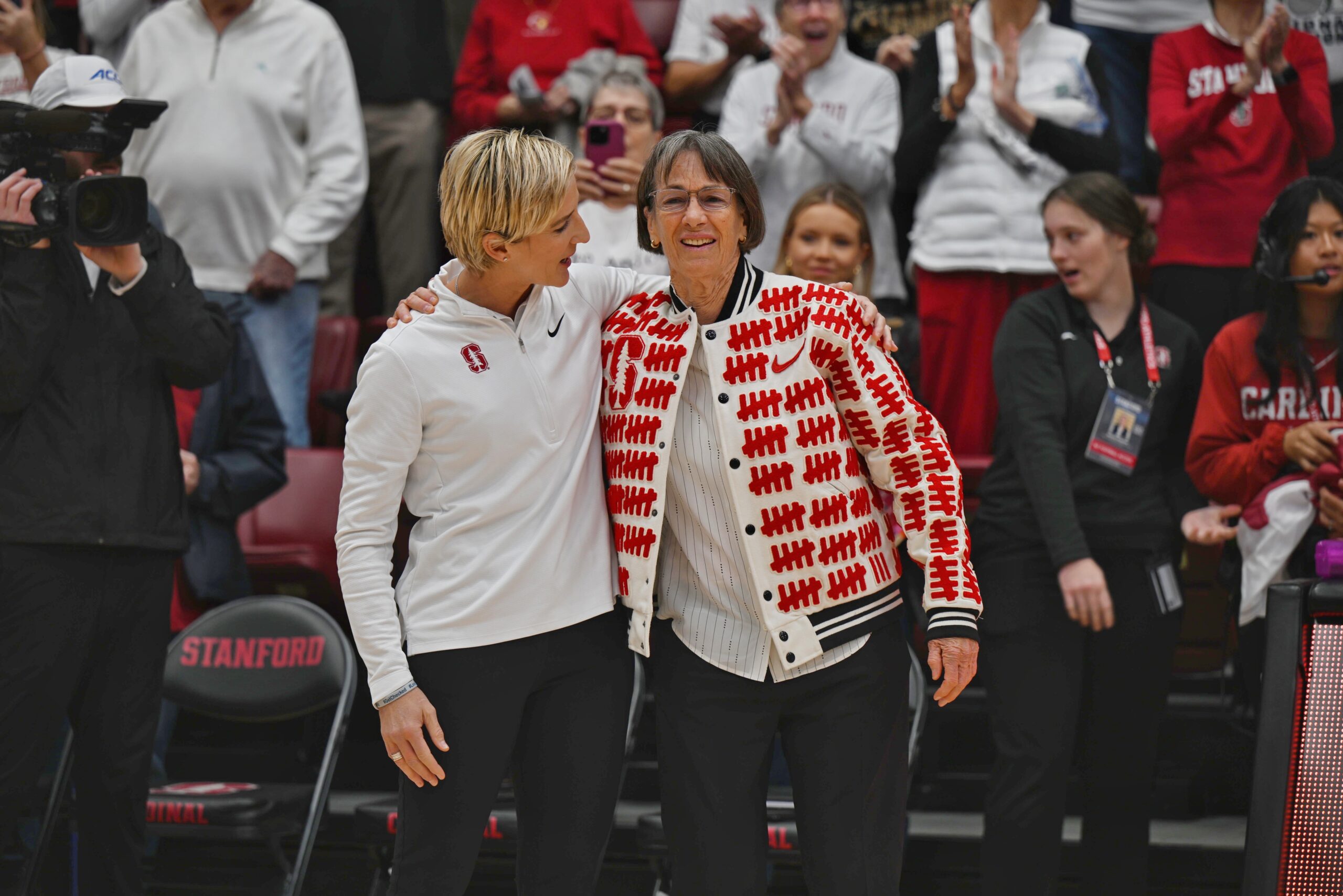 Stanford head coach Kate Paye and retired coach Tara VanDerveer stand together arm in arm at Maples Pavilion.