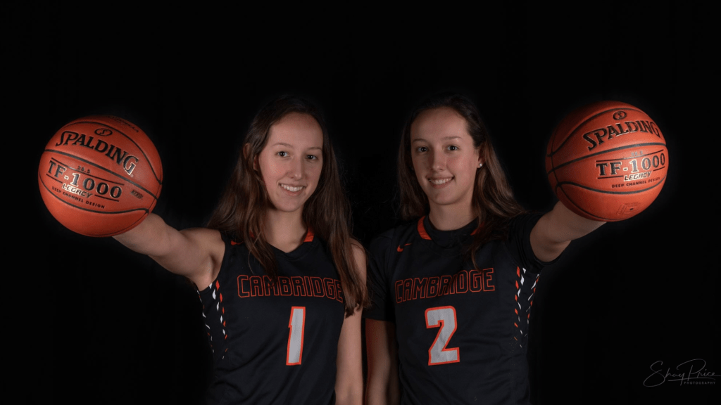 Lilly (1, left) holds her right arm out straight with a Spalding basketball in her hand. Next to her, sister Sophie (2, right) holds her left arm straight out in from of her while holding an identical Spalding basketball. Both are smiling and facing the camera.