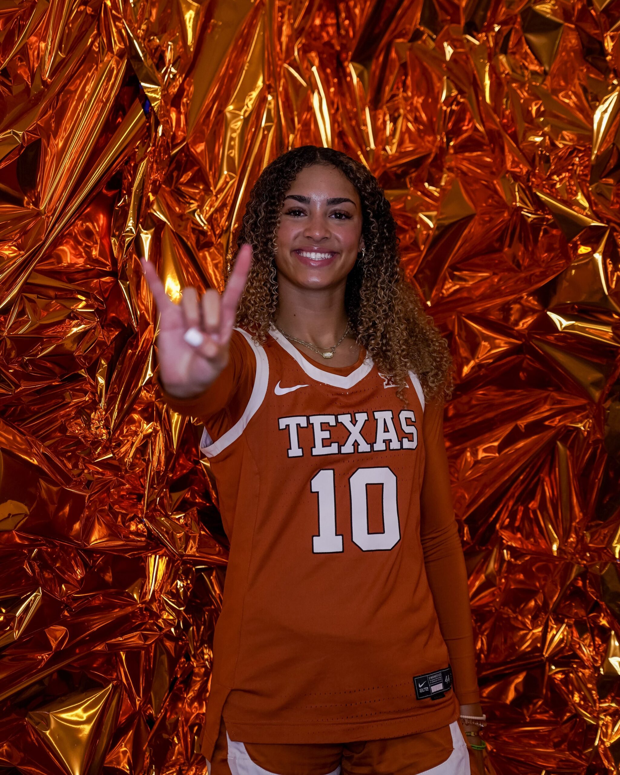 Photo of incoming Texas guard Aaliyah Crump smiling and holding up a Longhorns hand sign in front of a shiny textured orange background.