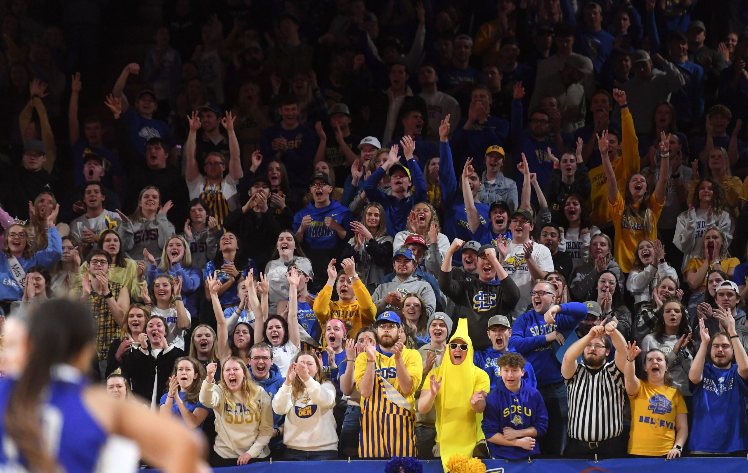 A fully packed crowd of South Dakota State fans stands cheering, clapping and/or holding up three fingers in celebration.