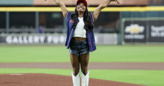 Simone Biles throwing out a first pitch at a Houston Texans Game, throwing her hands in the air.