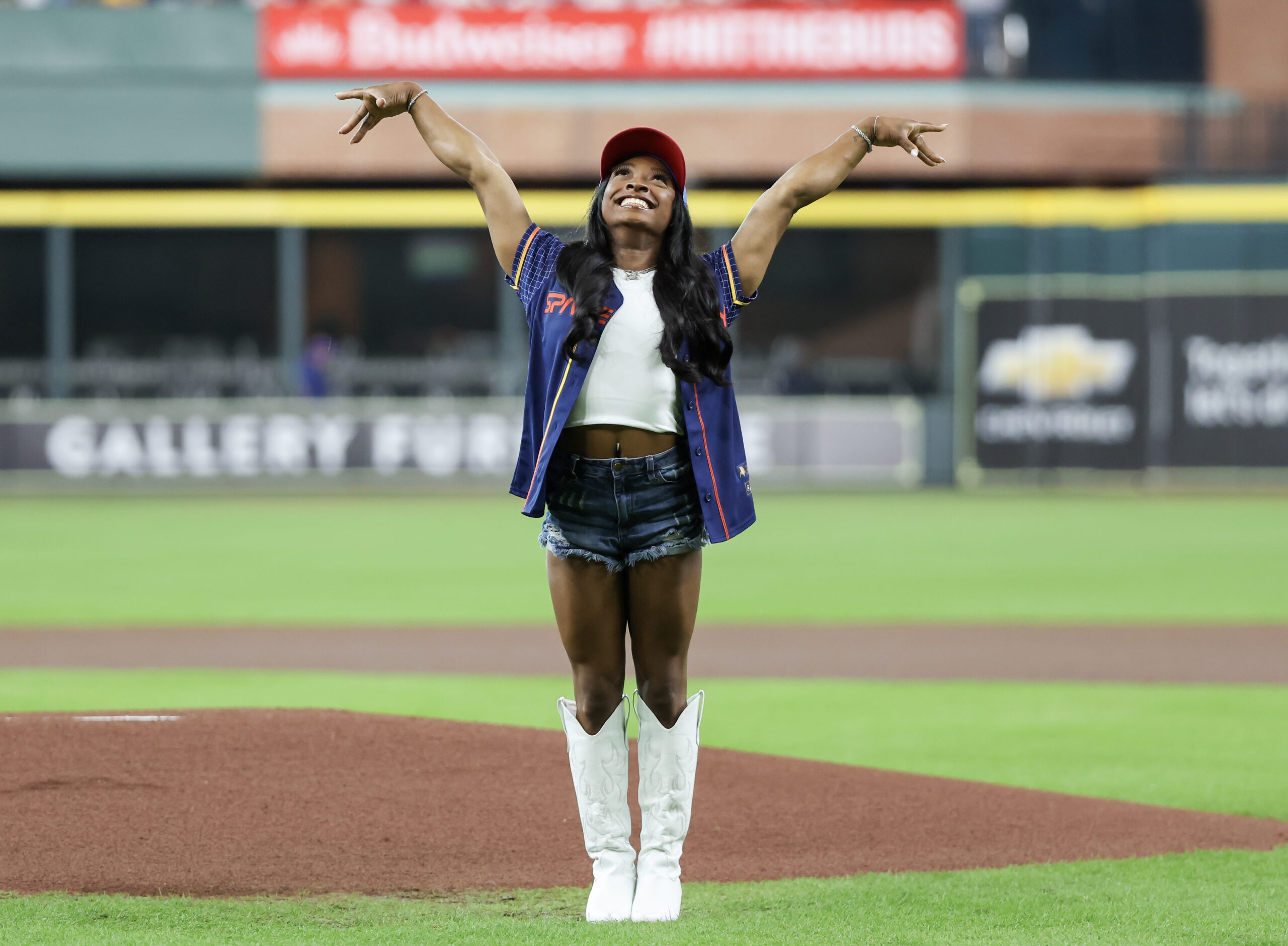 Simone Biles throwing out a first pitch at a Houston Texans Game, throwing her hands in the air.