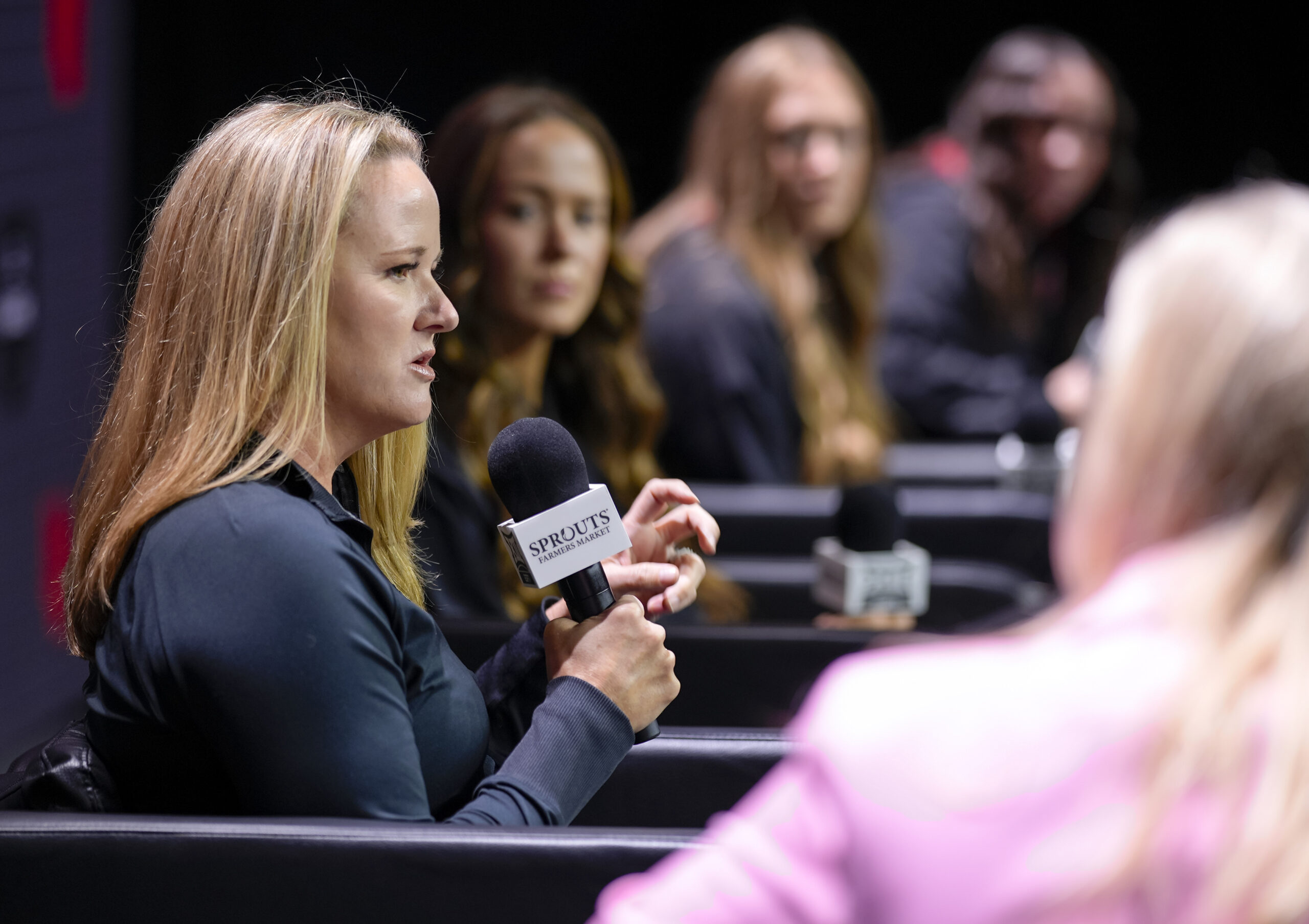 Utah head coach Lynne Roberts is shown in profile as she answers questions from reporters at Big 12 media day. She is seated in an armchair and holds a microphone in her right hand.