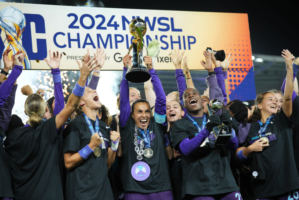 Photo of Marta holding the NWSL Championship trophy up in the air. She is joined on stage by her Orlando Pride teammates. Everyone is smiling and celebrating.