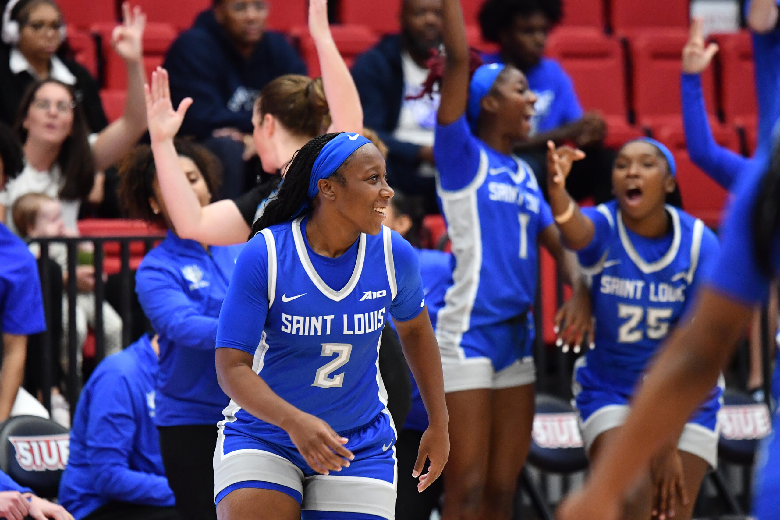 Saint Louis guard Shun'teria Anumele looks to her left and smiles as fans and a teammate in the background hold up three fingers to signal a made 3-pointer.