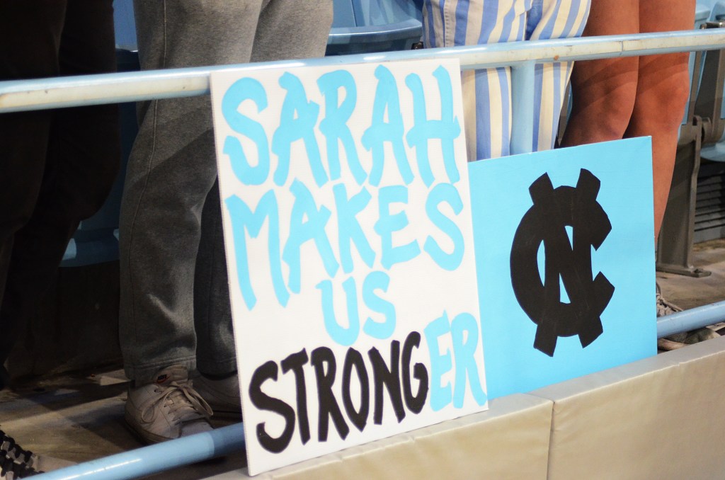 Two signs lean against a railing at a UNC game. At left, a poster with a white background with blue text "Sarah Makes Us", black text "Strong" and blue text "er". At right, a blue background with the UNC logo in black at center.