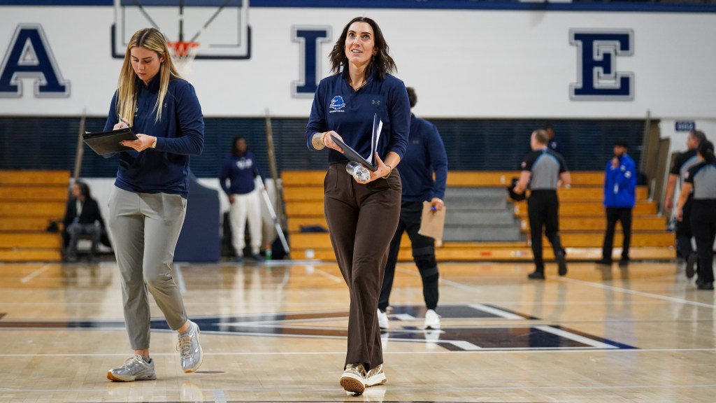 Monmouth head coach Cait Wetmore and director of player development and assistant coach Lola Mullaney walk across the court. Wetmore looks up and holds a half-open folder. Mullaney writes something down as she walks.