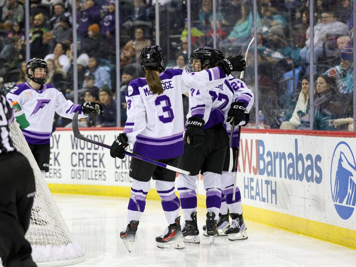Four Frost players celebrate with a group hug behind the net. They are wearing white away uniforms.