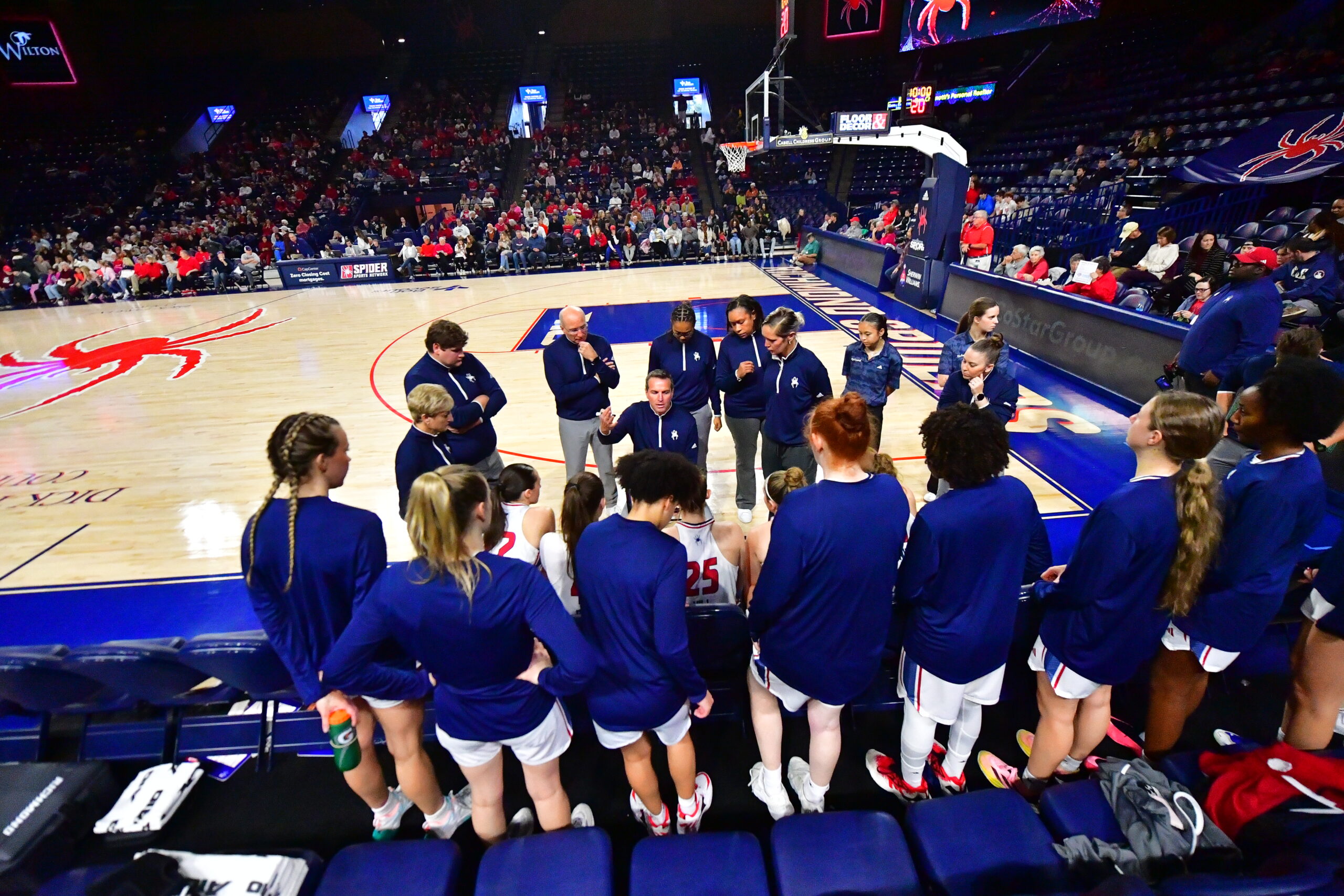 Richmond players sit and stand while looking at head coach Aaron Roussell who is leading the huddle with a marker in his hand. Roussell's coaching staff stand behind him, listening intently.